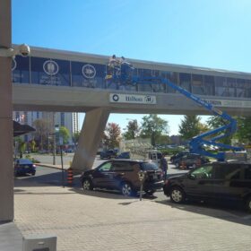 Installers using boom lift to install window graphics on bridge connecting the Hilton to the RBC place